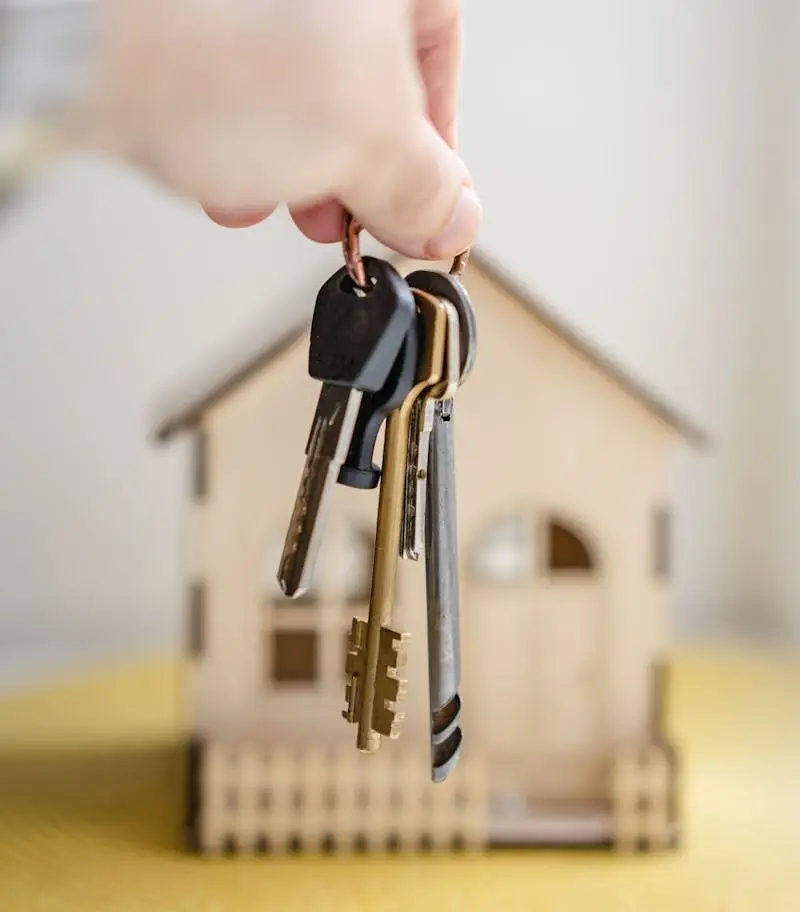Close-up of a hand holding keys with a miniature wooden house in the background, symbolizing real estate investment.